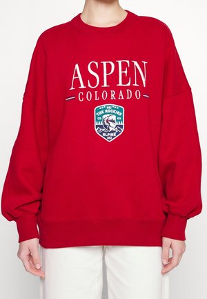 Sudadera roja de gran tamaño con "ASPEN COLORADO" impreso en blanco. Presenta una insignia de esquí en verde y negro en la parte frontal. Tejido suave y texturizado.