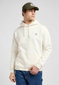 Sudadera color crema con un bolsillo tipo canguro, capucha con cordón y un pequeño parche con logo. Llevada con jeans azules y una gorra verde. Textura suave.