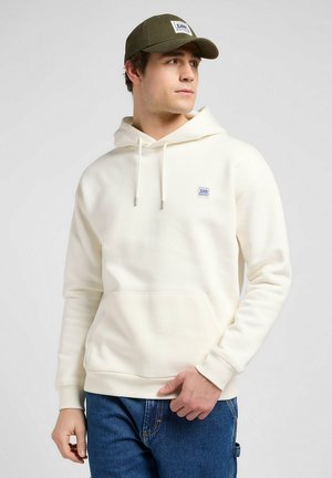 Cream-colored hoodie with a kangaroo pocket, drawstring hood, and a small logo patch. Worn with blue jeans and a green cap. Smooth texture.