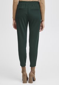 Vrouw in donkere groene, taps toelopende broek met elastische tailleband, een beige langemouwtop en bijpassende, hoge enkellaarsjes, gezien van achteren.