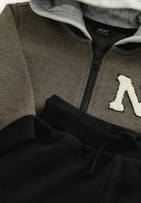 Huppari, jossa on ruskea ruudullinen kuvio ja mustat resorit, yhdistetty mustiin collegehousuihin. Huomattava valkoinen fleecestä valmistettu "N" -embleemi päällä.