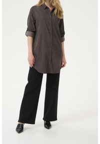 Langærmet, skjorte med knapper i mørkebrunt fløjlsstof, med brystlomme, kombineret med vide sorte bukser og spidse sorte sko.