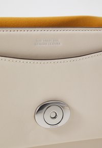 Intérieur d'un sac en cuir crème avec une doublure en daim moutarde douce. Comprend un accent de quincaillerie circulaire en argent et un détail de texte en relief.