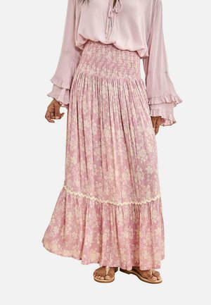 Mujer con falda larga rosa con estampado floral, cintura fruncida y bajo con volantes, combinada con una blusa rosa clara de mangas acampanadas y sandalias.