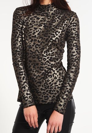 Kvinde iført langærmet, metallisk leopardprint-top kombineret med sorte bukser, stående mod en ensfarvet hvid baggrund.