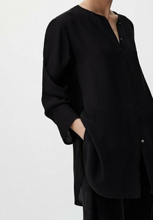 Femme portant un chemisier noir ample à manches longues avec col mandarin et deux boutons, une main dans la poche, sur un fond clair uni.