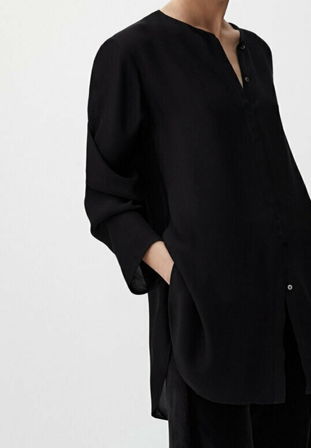 Femme portant un chemisier noir ample à manches longues avec col mandarin et deux boutons, une main dans la poche, sur un fond clair uni.