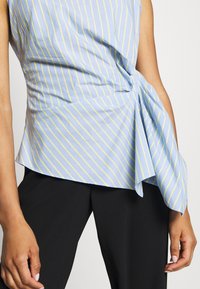 Ljusblå ärmlös topp med gula vertikala ränder, med en vriden frontdetalj och en utställd nederkant, i kombination med svarta byxor.