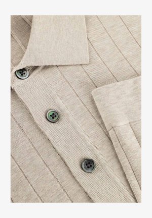 Beige geribbelde gebreide cardigan met een kraag, drie zichtbare donkere knopen en een omgeslagen manchetknoop aan de mouw.