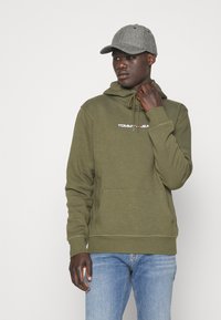 Sudadera color verde oliva con bolsillo frontal y capucha ajustable, que presenta el logo "TOMMY JEANS" en blanco. Combinada con jeans azul claro y una gorra gris.