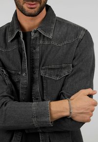 Giubbotto di denim nero con bottoni a pressione, due tasche sul petto e una finitura testurizzata. Un bracciale in metallo argentato adorna il polso.