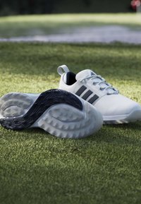 Λευκά παπούτσια του γκολφ με μαύρες σόλες και τρεις μαύρες ρίγες πάνω σε γρασίδι κοντά σε έναν αμμόλοφο με μια κόκκινη σημαία στο βάθος.