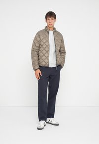 Gepolsterte, beige Pufferjacke mit hohem Kragen, kombiniert mit einem hellgrauen Sweatshirt und marineblauen Hosen. Weiße Sneakers mit schwarzen Akzenten.