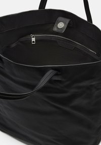 Sac fourre-tout en cuir noir avec une texture lisse, une poche zippée et un fermoir à pression. L'intérieur est doublé de tissu noir. Dispose de poignées robustes.