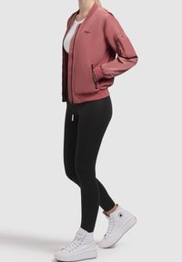 Femme portant une veste bomber rose poussiére, un haut blanc, des leggings noirs et des baskets Converse montantes blanches, debout de profil.