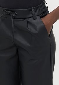 Pantalons en simili cuir noir avec une taille élastique, détails de cordon de serrage, poches latérales et plis. Texture lisse et coupe ajustée.