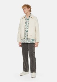 Cream-colored jacket with a collar, layered over a green tropical print shirt. Dark grey cargo pants and white sneakers complete the outfit.