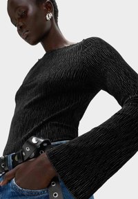 Czarny strukturalny top z długim rękawem o falistym wzorze, z rozkloszowanymi rękawami. Noszony z niebieskimi dżinsami i czarnym paskiem z ćwiekami.
