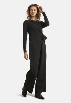 Schwarzer Jumpsuit mit langen Ärmeln und Wickeldesign. Mit einer gebundenen Detail an der Taille, fließenden weiten Beinen und eleganten schwarzen High Heels.