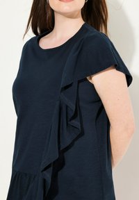 Granatowa bluzka z krótkim rękawem i okrągłym dekoltem, z falbanką warstwową wzdłuż ramienia oraz luźnym, strukturalnym krojem.