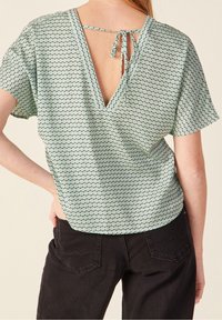 Donna che indossa una blusa a maniche corte con un motivo geometrico verde menta, con schiena scoperta e chiusura a cravatta, abbinata a jeans neri.
