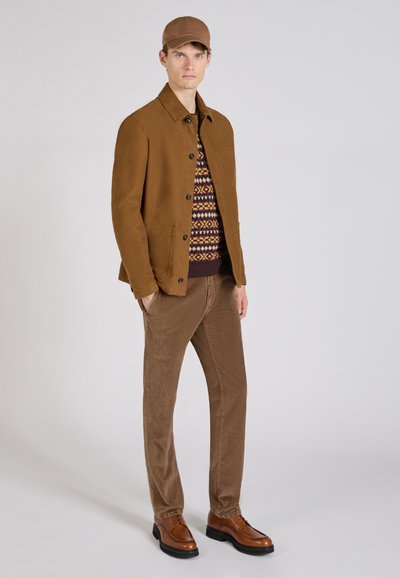 Braune Wildlederjacke über einem gemusterten Pullover, beige Cordhosen und braunen Lederschuhen. Die Accessoires umfassen eine passende braune Mütze.