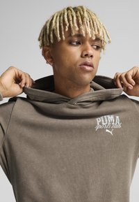 Sudadera marrón con un gran cuello doblado. Presenta un logo bordado de "PUMA sports club" y una textura suave. Mano sobre el cuello.