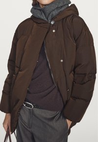 Brun puffet jakke med hætte, med lynlås foran og trykknapper, båret over en mørk sweater og kombineret med grå bukser og et sort bælte.