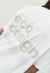 Biela tričko s ekologickými dizajnovými princípmi v svetlošedej farbe. Tkanina vyzerá mäkká, s voľným strihom a minimalistickým dizajnom.