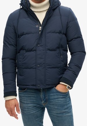 Homme portant une veste matelassée bleu marine par-dessus un pull à col roulé blanc et un jean bleu, une main dans la poche et une montre-bracelet visible.