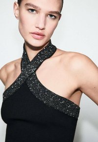 Top de punto negro con diseño de cuello halter. El escote está adornado con lentejuelas negras, creando un efecto texturizado y embellecido.