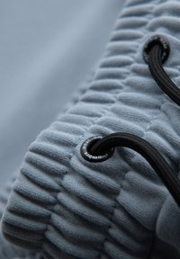 Tissu bleu clair avec un design plissé texturé. Présente un cordon noir passé à travers un œillet rond avec une marque.