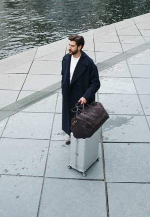 Silver aluminum suitcase with wheels, paired with a dark brown leather duffel bag. Gray textured pavement and water in the background.