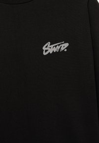 Tissu noir avec un petit logo texte stylisé gris clair imprimé sur la partie supérieure de la poitrine.