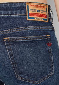 Tmavě modré džíny z denimu s viditelným šitím, s označením kožené značky na zadní straně a malým červeným logem na kapse.