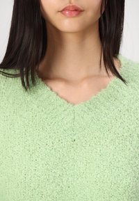 Pull en maille vert clair et duveteux avec un col en V, présentant une surface texturée et une coupe ample. Gros plan sur la zone de la poitrine.