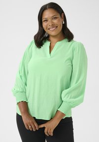 Blouse vert menthe avec une coupe décontractée, texture douce et manches bouffantes plissées. Présente un décolleté en V et des poignets smockés, portée avec un pantalon noir.