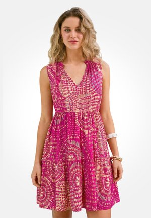 Žena v ružových šatách bez rukávov s krémovými geometrickými vzormi, ozdobená náramkami na pravom zápästí, stojaca na bielom pozadí.