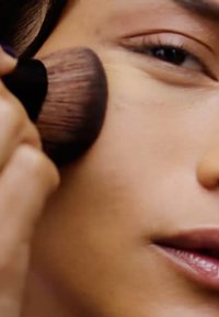 Un pinceau à maquillage appliquant du fond de teint sur la peau. Le pinceau possède un manche en bois et des poils doux ; la peau paraît lisse et uniformément teintée.