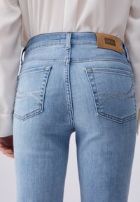 Blugi bleu deschis cu detaliu din strasuri pe buzunarele din spate, purtați cu o cămașă albă cu mâneci lungi, văzuți din spate, la nivelul taliei.