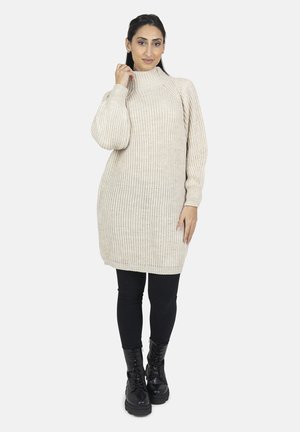 Beige strikket kjole med høj krave, ribbet struktur, lange ærmer og løs pasform, parret med sorte leggings og kraftige sorte støvler.