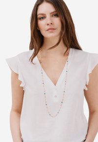 Blusa bianca in lino con maniche corte a volant, profondo scollo a V e tre bottoni. Collana di perline con accenti colorati nei toni pastello e acquamarina.