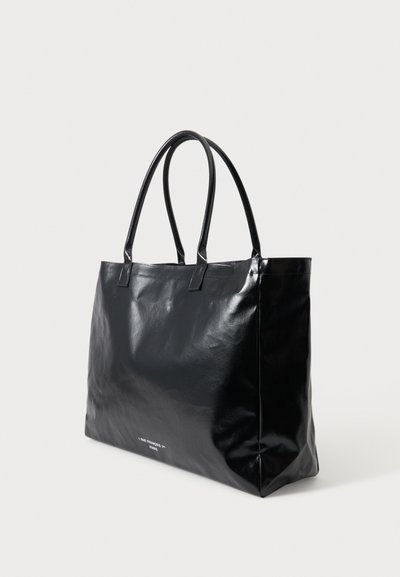 Sac cabas en cuir noir avec deux poignées et petit texte blanc sur le devant, posé devant un fond clair uni.