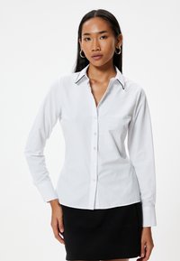 Camisa blanca de manga larga con cuello, que presenta un ribete negro en contraste y cierre de botones. Textura suave, diseño ajustado.