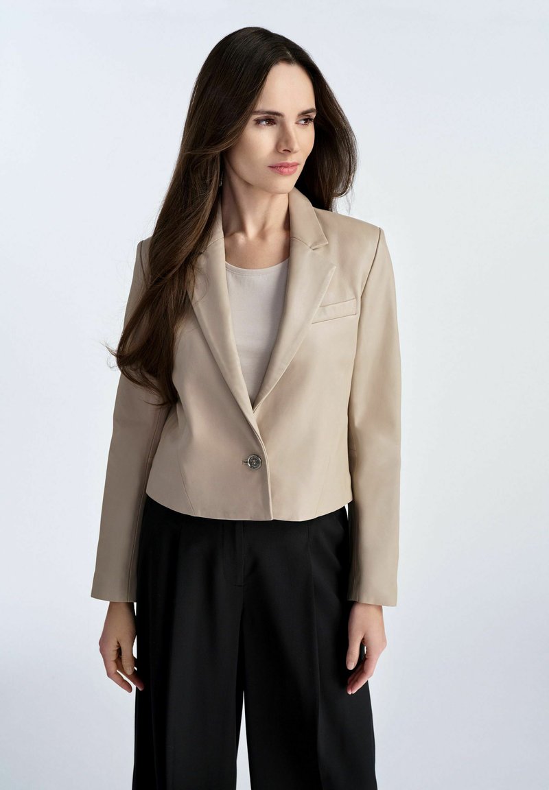 Beżowa krótka marynarka uszyta z gładkiego materiału, z pojedynczym zapięciem na guzik i subtelną kieszonką, w zestawie z czarnymi spodniami.