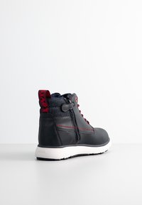 Baskets montantes noires avec coutures rouges, fermeture éclair sur le côté, languette rouge et semelle blanche sur un fond uni.