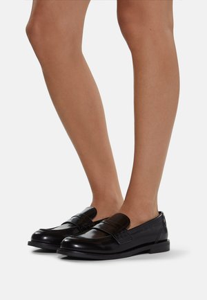 Piernas que llevan mocasines de cuero negro con tacones bajos, mostradas contra un fondo claro y liso.
