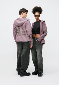 Purple hoodie with graphic print, paired with black knotted top, gray baggy jeans, and chunky black boots. Both figures face slightly backward.
