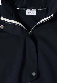 Veste marine avec une fermeture éclair à l'avant et des boutons-pression. Dotée d'une capuche doublée blanche et d'un tissu subtilement texturé ; l'étiquette indique "CECIL".