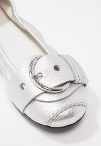 Chaussure plate en cuir texturé argenté avec une grande boucle argentée et un détail de couture arrondi à l'avant.